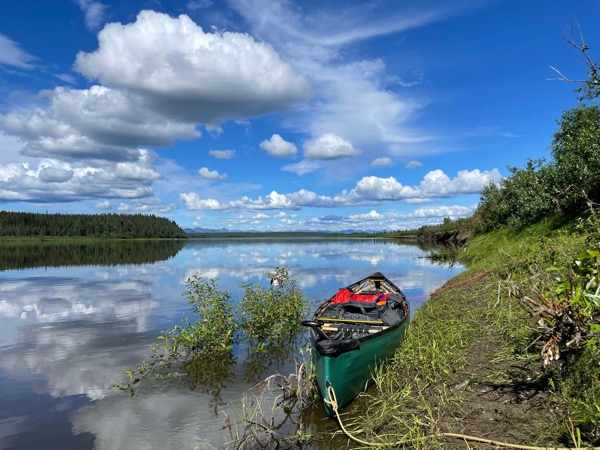 A green pack canoe sits on the bank of the Kobuk River. Clouds are mirrored on the water's surface.