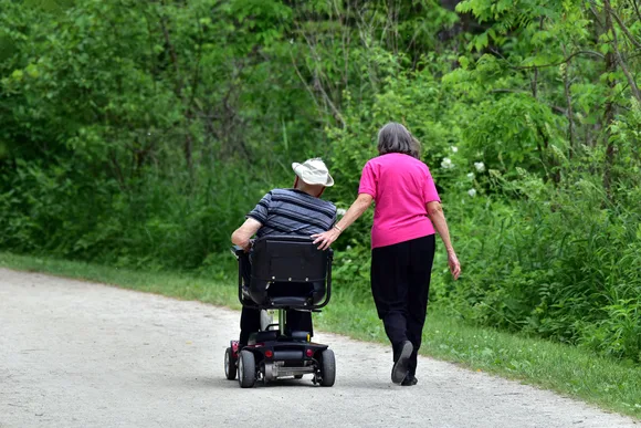 A woman walks beside a man on a flat trail, resting a hand on the back of his motorized wheelchair.