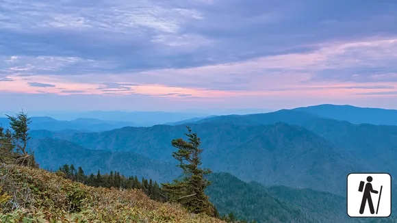 Sweeping mountain views under a pink and purple sky. Hiker icon in corner.
