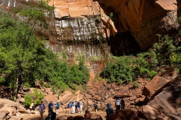 A large group of people near a pool of water with green trees and an orange sandstone cliff face