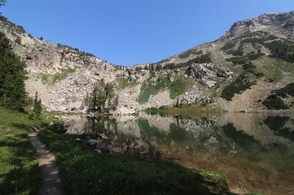 An alpine lake sits at the base of a rocky cliff surrounded by green vegetation.