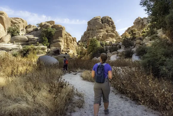 wo hikers walk along a sandy trail surrounded by golden vegetation and tall granite rock formations