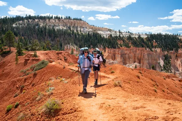 Two people walk a trail of red earth wearing gear on their backs.